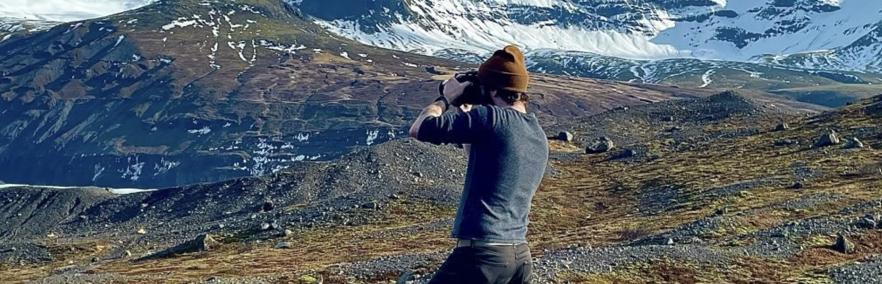 A person standing outdoors on rocky terrain with snow-capped mountains in the background, wearing a brown beanie, sunglasses, and a long-sleeve shirt.