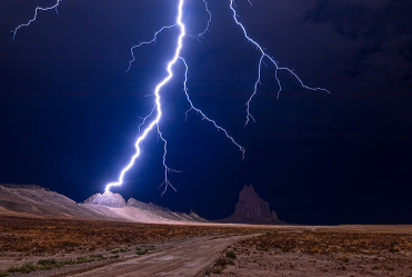 Lightning strikes over a desert landscape with dark stormy skies and distant mountains.