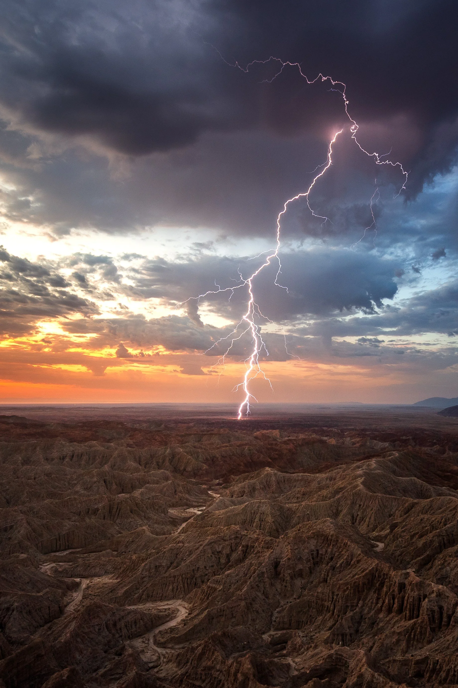 Lightning storm over a rugged, arid landscape at sunset with dark clouds overhead.