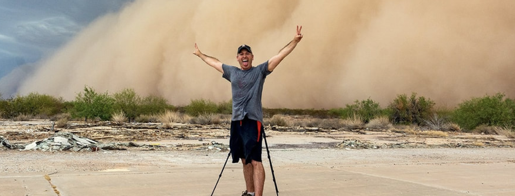 A man smiling with arms raised making peace signs standing on a desert road with a dust storm and sparse desert vegetation in the background.