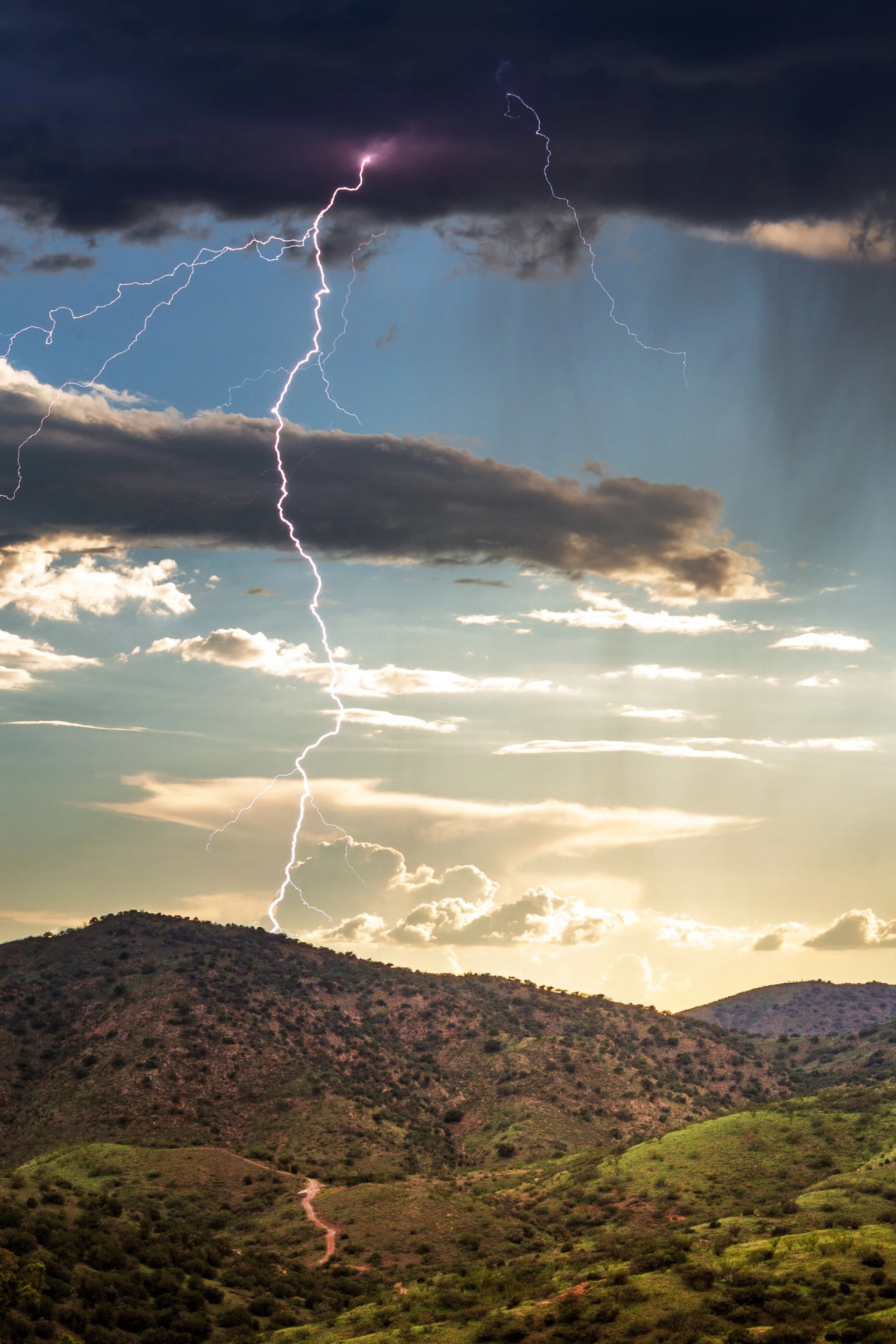 Lightning strikes in a cloudy sky over a green hilly landscape during a thunderstorm.