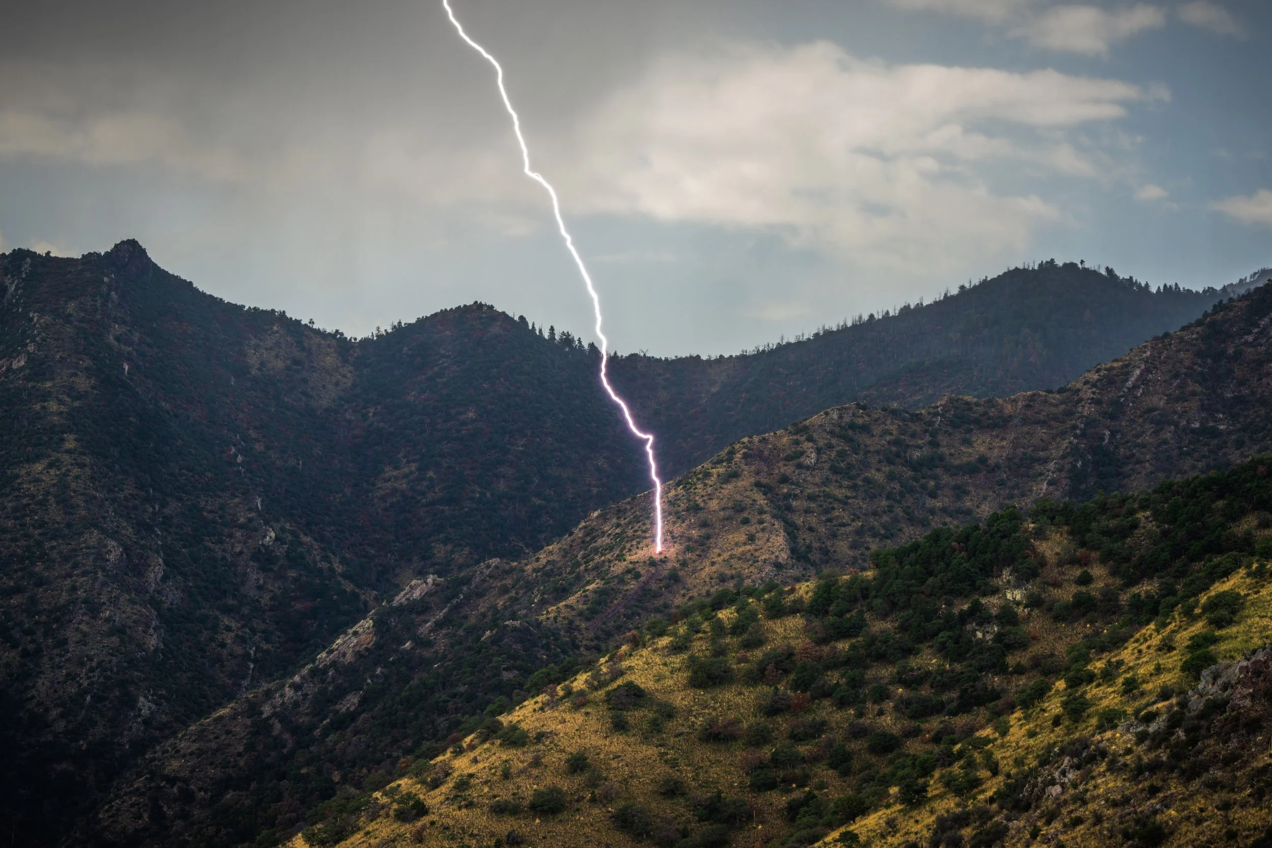 A lightning bolt strikes in the mountainous terrain on a cloudy day.