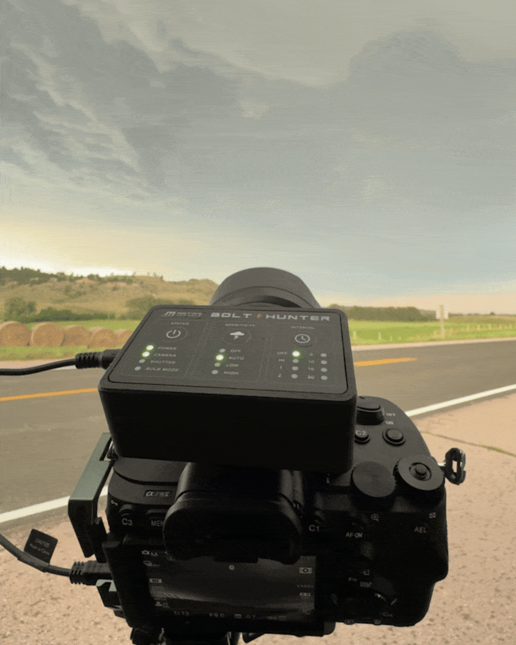 Camera mounted on a vehicle capturing an outdoor landscape of a roadside with hay bales, a hill, and a partly cloudy sky during sunrise or sunset.