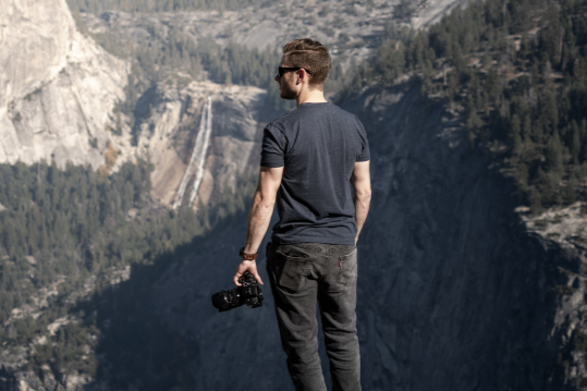 Man standing with his back facing a mountain valley, holding a camera, wearing sunglasses and casual clothing.