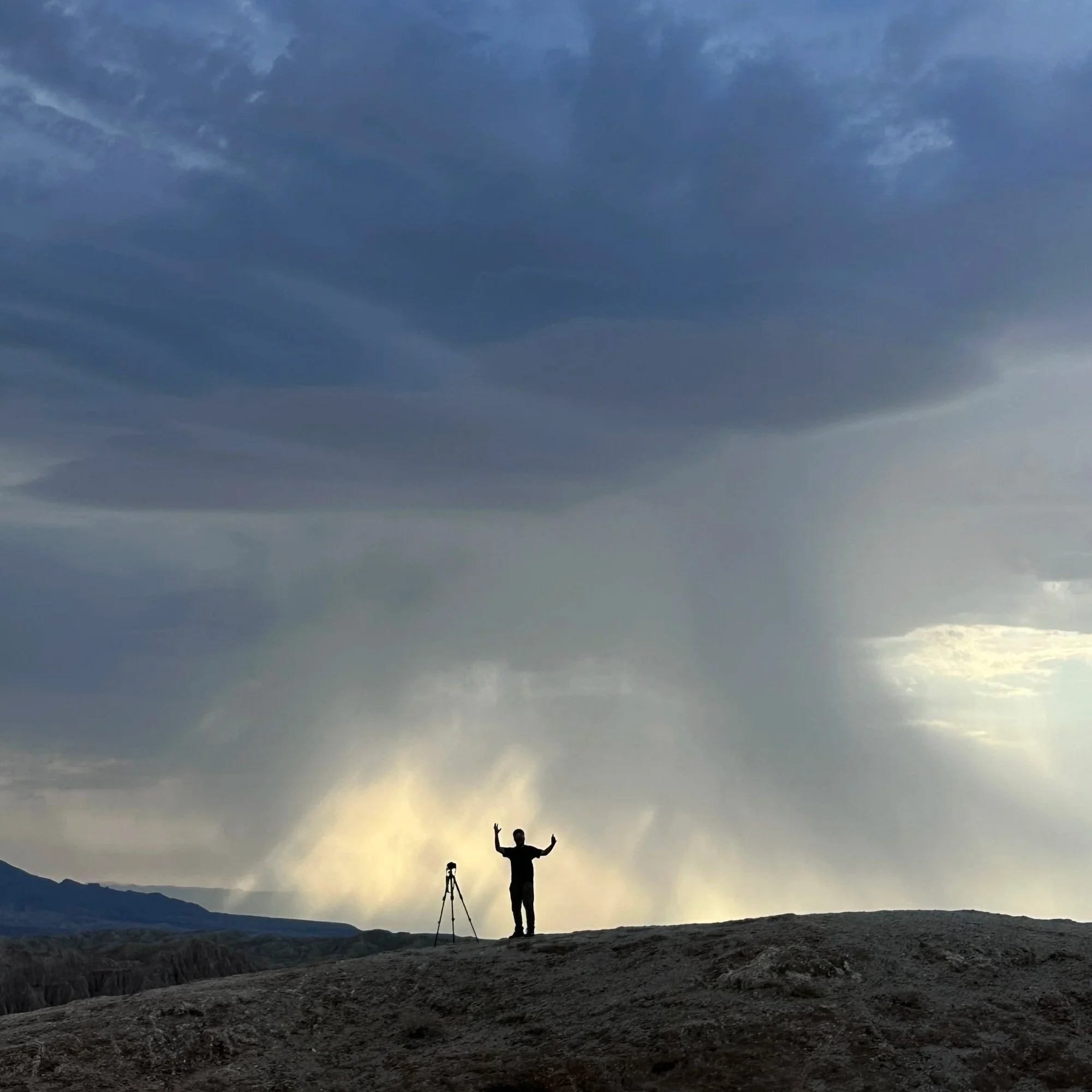 Person standing on rocky terrain with arms raised, near a tripod, under a dramatic sky with dark clouds and rain in the distance.