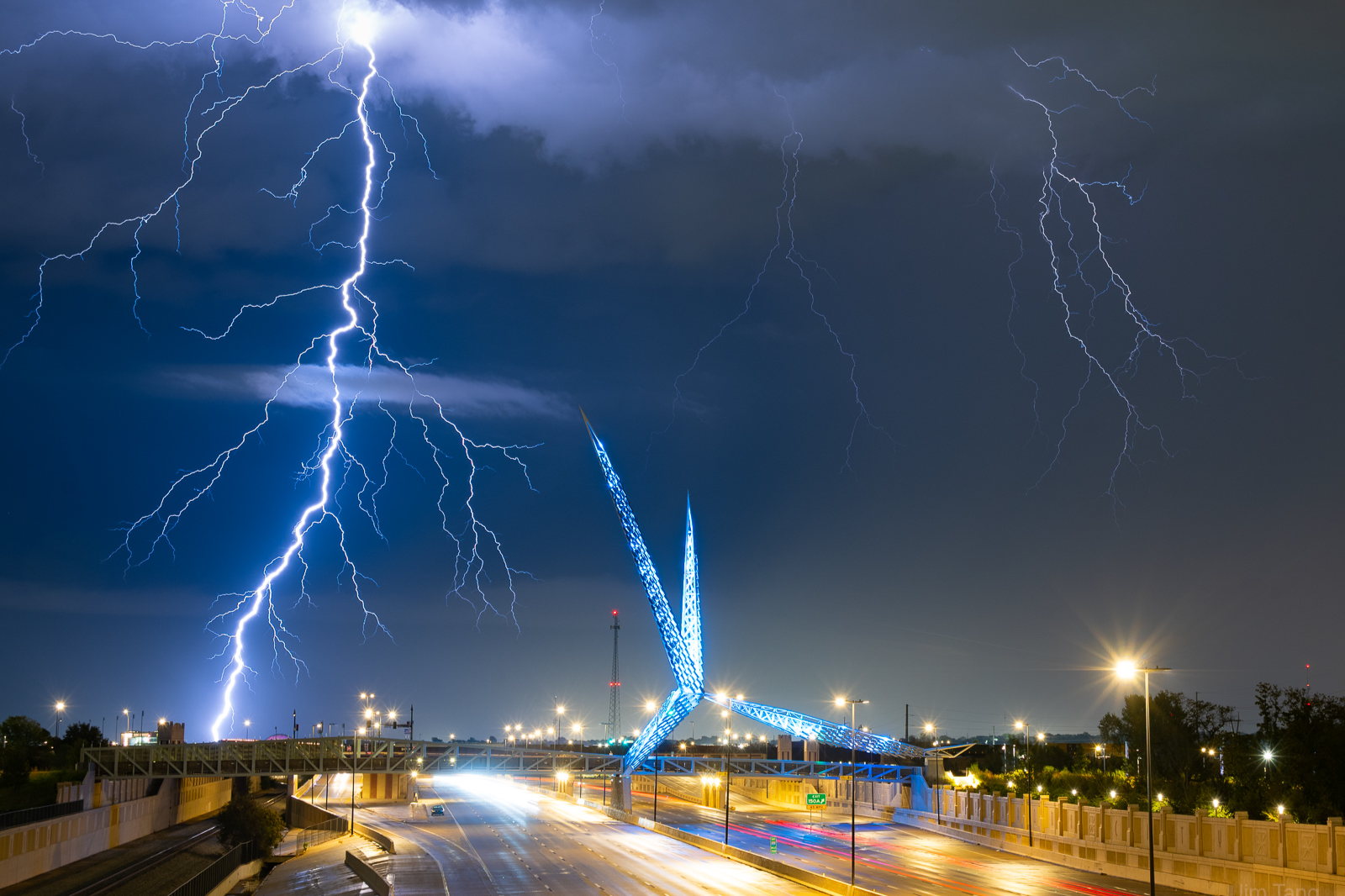 Lightning strikes over a bridge with a large, illuminated blue sculpture in the shape of a bird, on a highway at night.