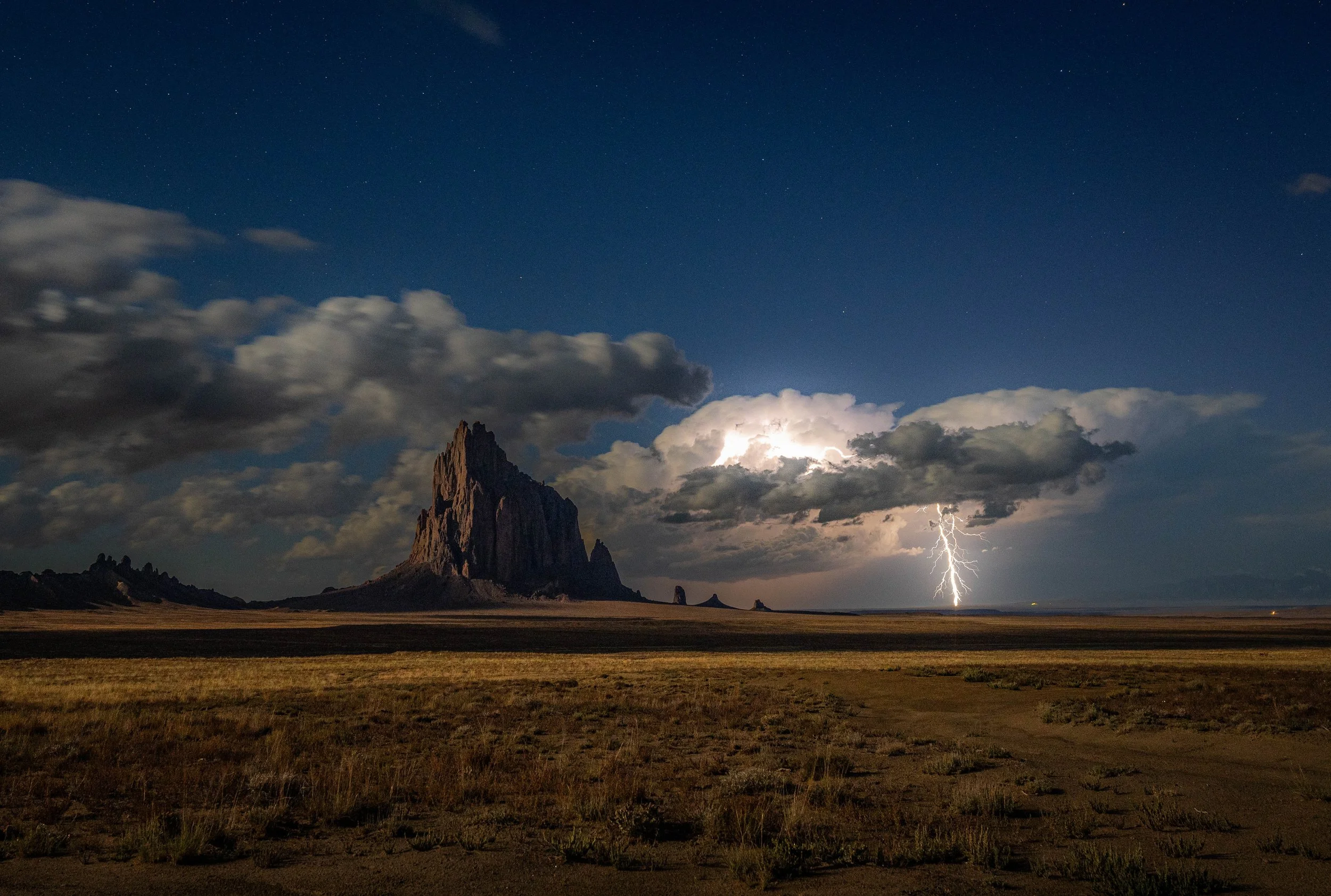 Thunderstorm over a flat desert landscape with a large mountain and lightning in the distance at night.