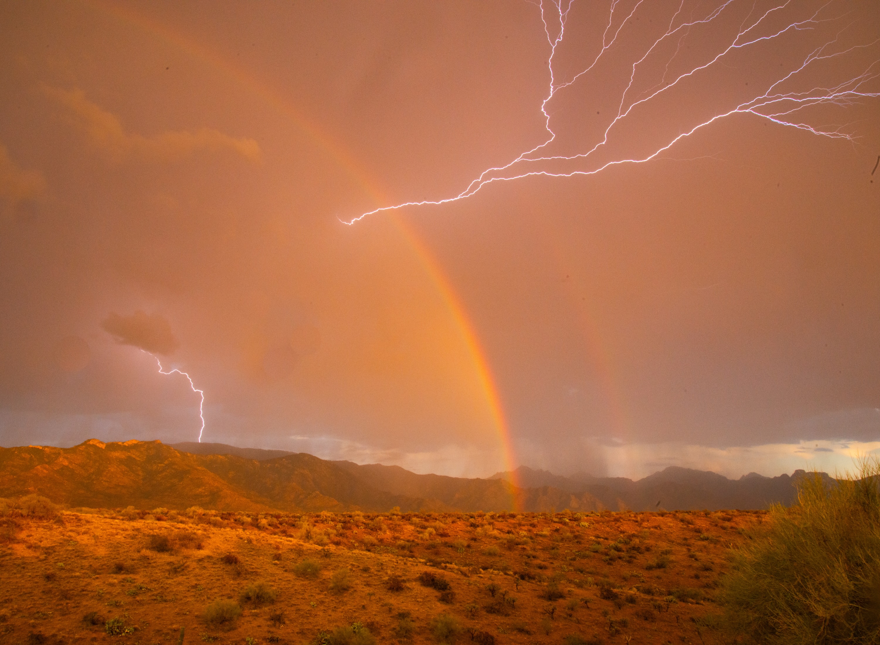 Lightning crossing an orange rainbow over the desert